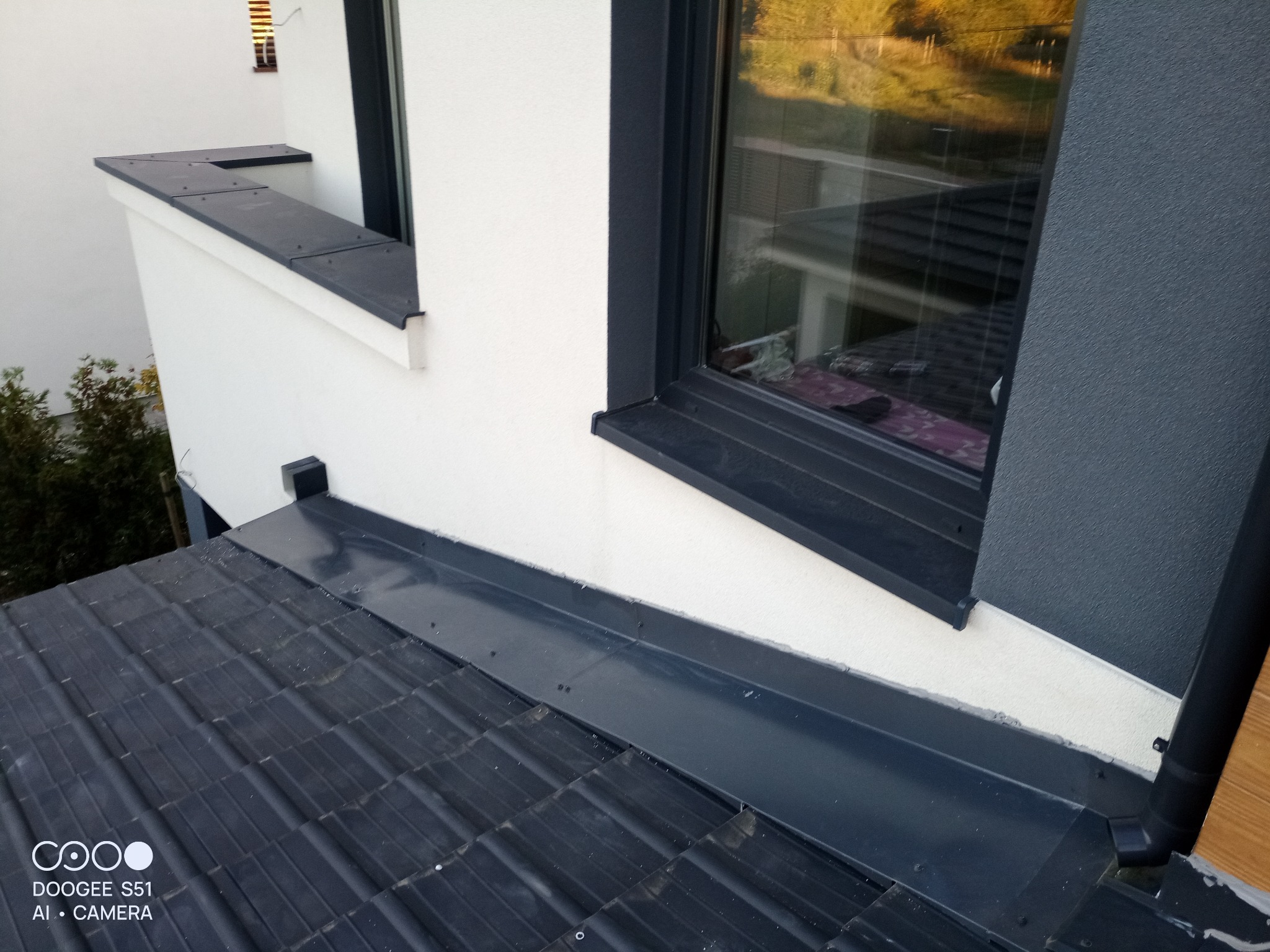 Nowe obróbki blacharskie okna i dachu w Pruszkowie. Ciemna blacha kontrastuje z jasną elewacją budynku. Widoczna rynna i dachówka.