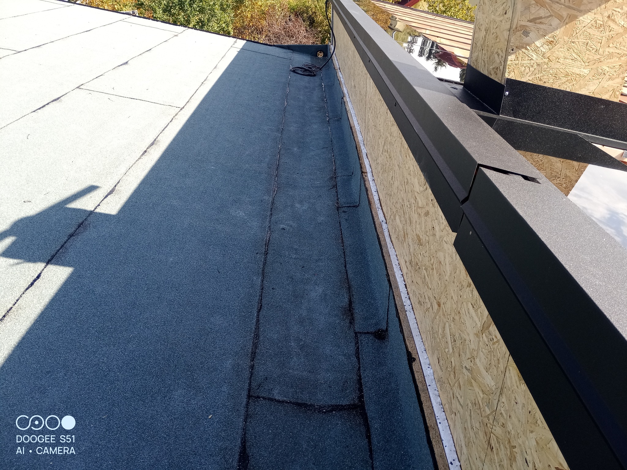 Fragment dachu pokrytego papą, z widocznymi obróbkami blacharskimi w kolorze czarnym i deskowaniem z płyty OSB. Widoczny kabel na dachu.