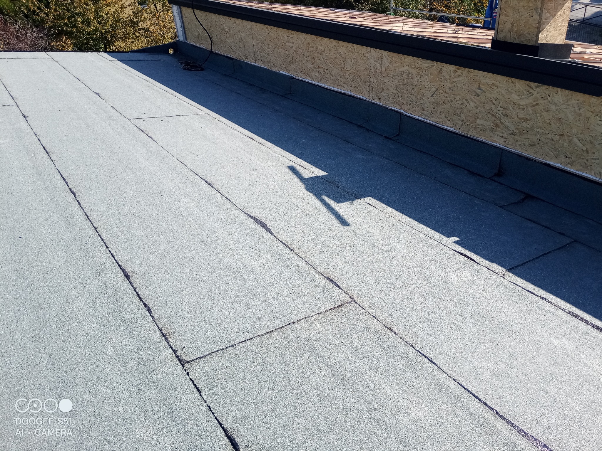 Szary, płaski dach pokryty papą termozgrzewalną z widocznymi łączeniami. W tle fragment ściany z płyt OSB i cień osoby.