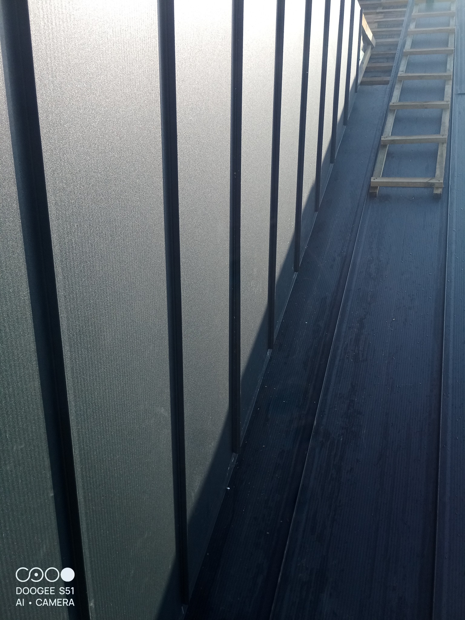 Szary dach z blachy trapezowej w pionowym ujęciu z drewnianą drabiną. Widoczne mocowania blachy. Dach oświetlony słońcem.