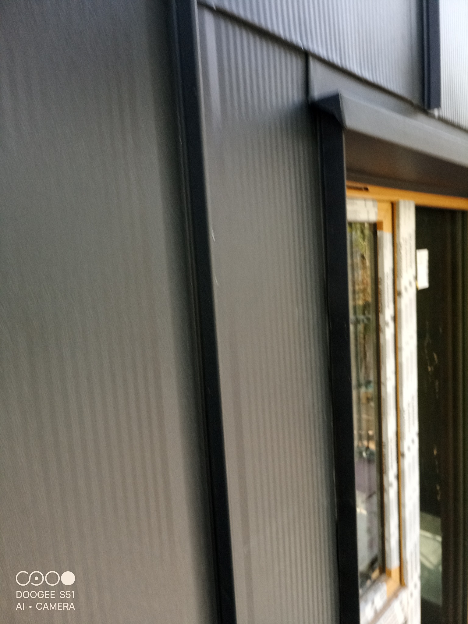 Szara, blaszana elewacja budynku z widocznym oknem w czarnej ramie. Pionowe przetłoczenia blachy. Ujęcie z dołu, podkreślające wysokość budynku.