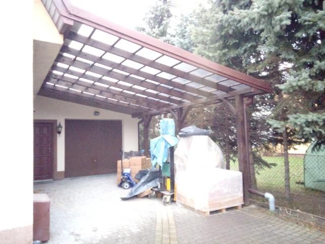 Drewniana wiata garażowa z przezroczystym zadaszeniem, przylegająca do budynku z dwoma bramami garażowymi. Częściowo zasłonięta materiałami budowlanymi.