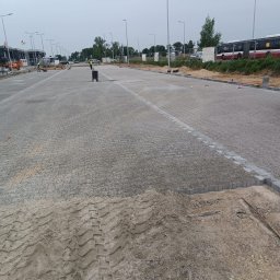 SVOT Viktor Stepanov - Rozległy plac wyłożony szarą kostką brukową w trakcie budowy, widoczne ślady opon na piasku i pracownicy w oddali, oświetlenie uliczne i autobusy w tle.