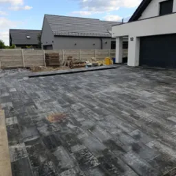 Nowoczesny podjazd z szarej, prostokątnej kostki brukowej, z widocznym włazem kanalizacyjnym i paletami kostki w tle, przy domu z numerem 12.