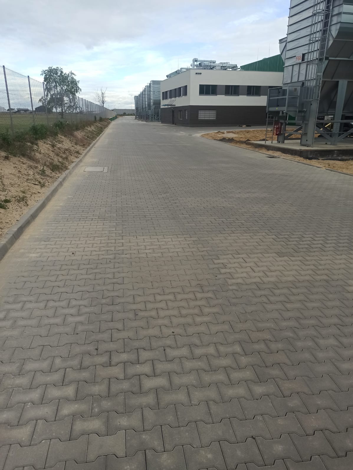Długa droga wyłożona szarą kostką brukową w kształcie litery H, prowadząca wzdłuż budynków przemysłowych z metalowymi silosami i ogrodzenia z siatki.