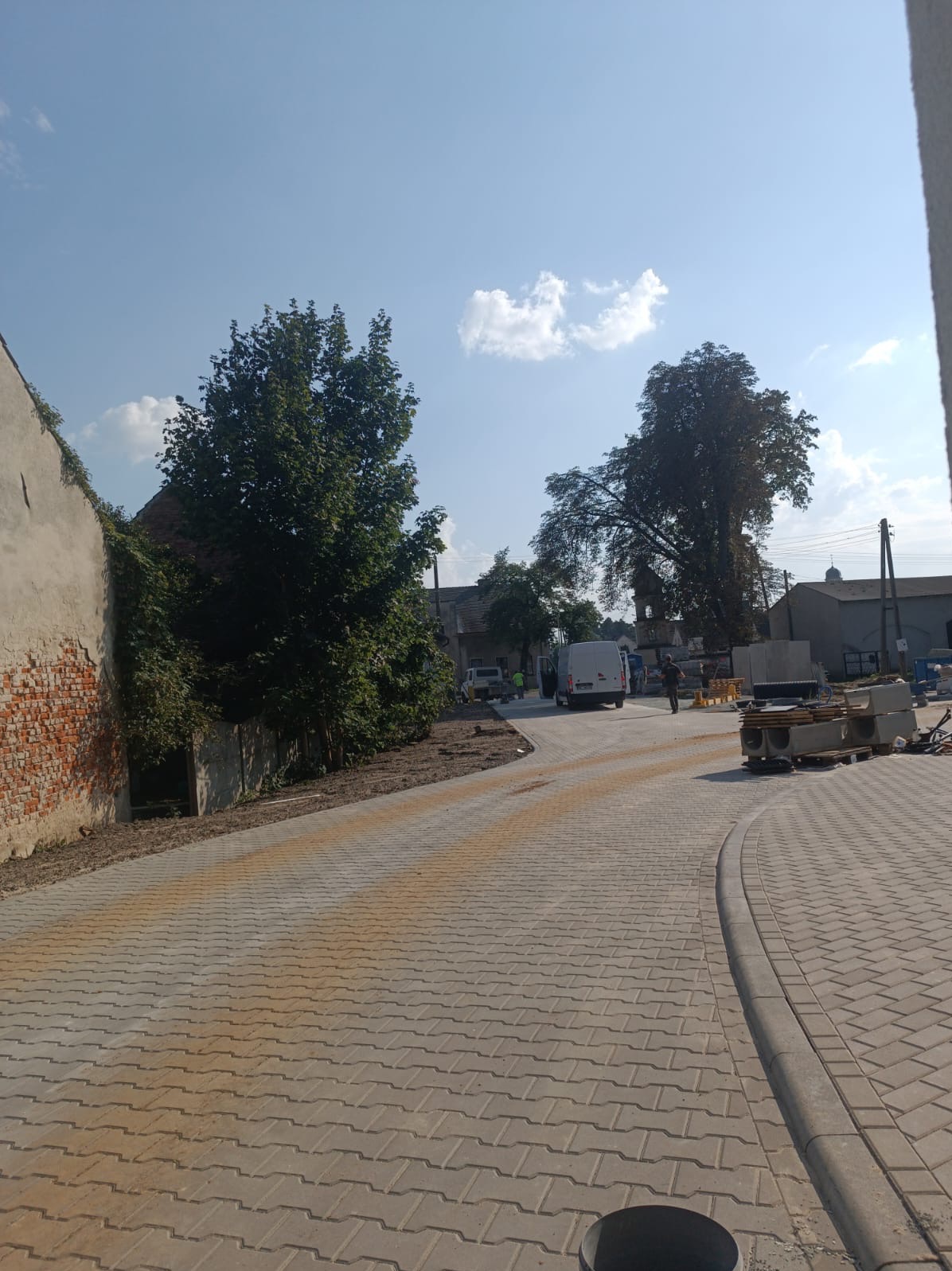 Nowo ułożona kostka brukowa w kształcie fali na drodze wiejskiej, z widocznymi śladami piasku, obrzeżami i zaparkowanym busem w tle, w słoneczny dzień.