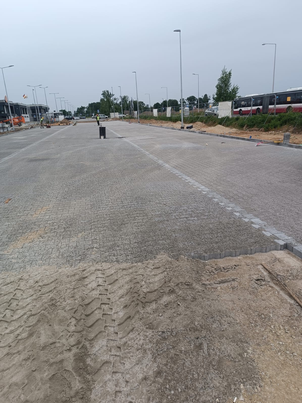 Rozległy plac wyłożony szarą kostką brukową w trakcie budowy, widoczne ślady opon na piasku i pracownicy w oddali, oświetlenie uliczne i autobusy w tle.
