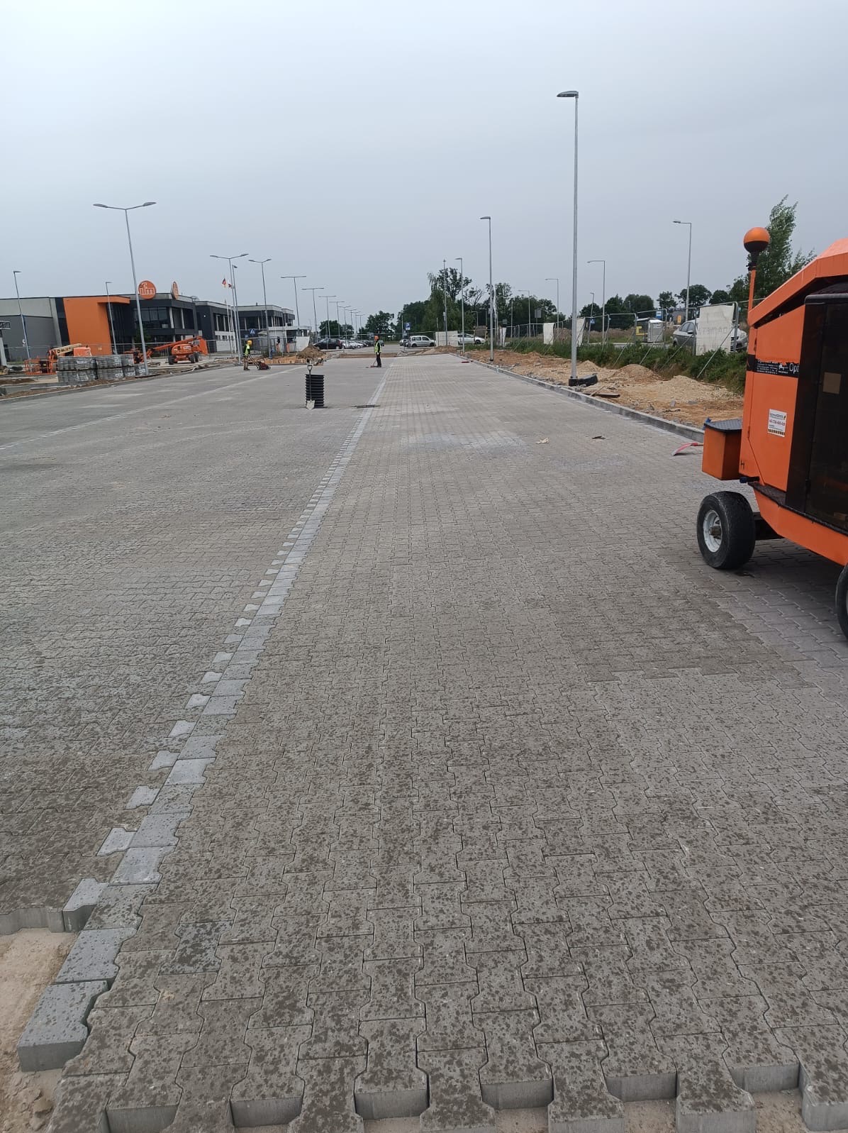 Rozległy plac wyłożony szarą kostką brukową typu 'puzzle', z widocznymi fragmentami piasku przy krawędziach, pomarańczowy podnośnik koszowy w tle po prawej stronie i budynki w oddali.