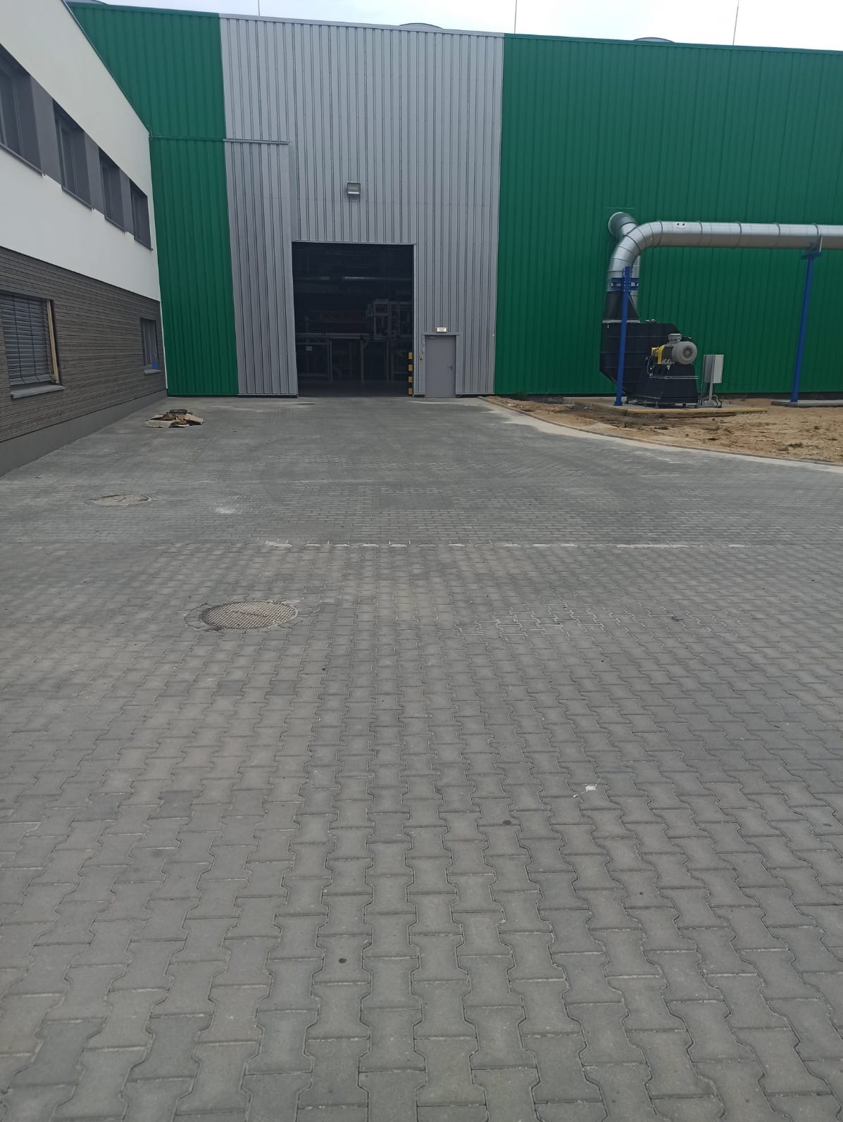 Szara kostka brukowa typu I na placu przed budynkiem przemysłowym z zielono-szarą elewacją, widoczne studzienki kanalizacyjne i instalacja wentylacyjna.