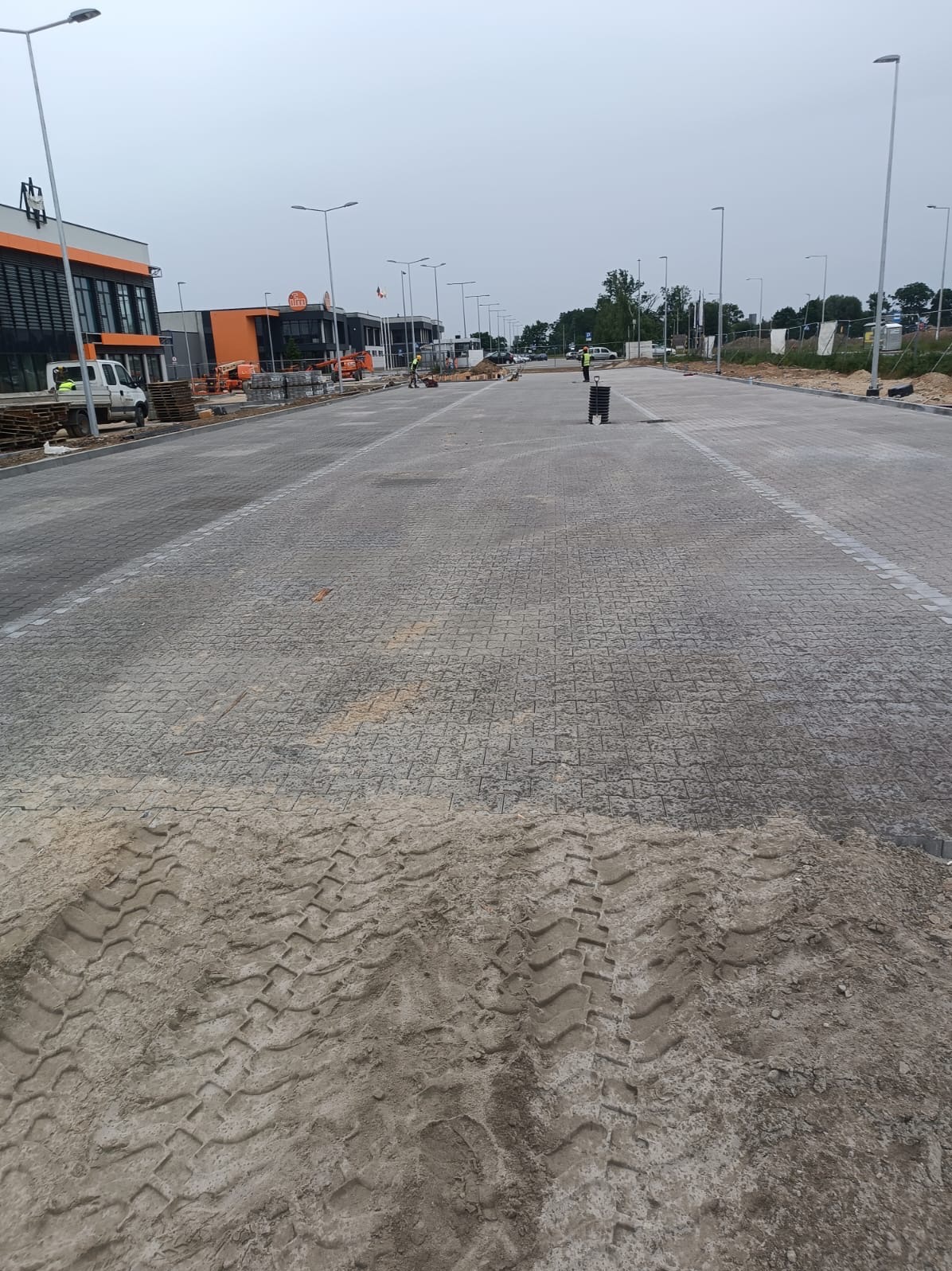 Rozległy plac wyłożony szarą kostką brukową w trakcie budowy, widoczne ślady opon na piasku, budynki w tle i latarnie.