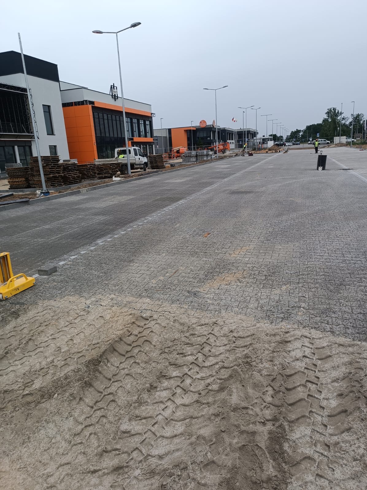 Szeroki kadr na plac budowy z ułożoną częścią szarej kostki brukowej, widoczne ślady opon na piasku w pierwszym planie, w tle budynki handlowe z elementami w kolorze pomarańczowym, palety z kostką...