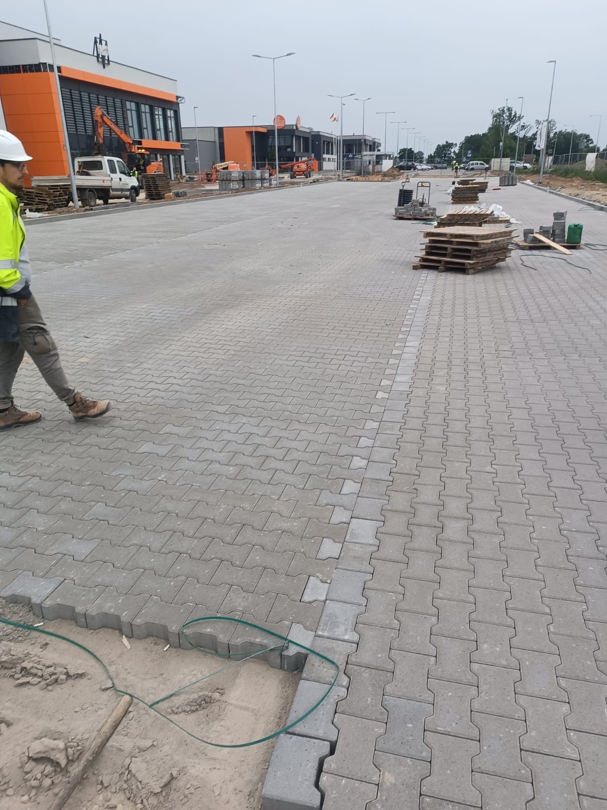 Układanie szarej kostki brukowej typu 'I' na dużej powierzchni placu, widoczny fragment wykopu, palety z kostką w tle, pracownik w odblaskowej kamizelce przechodzi obok.