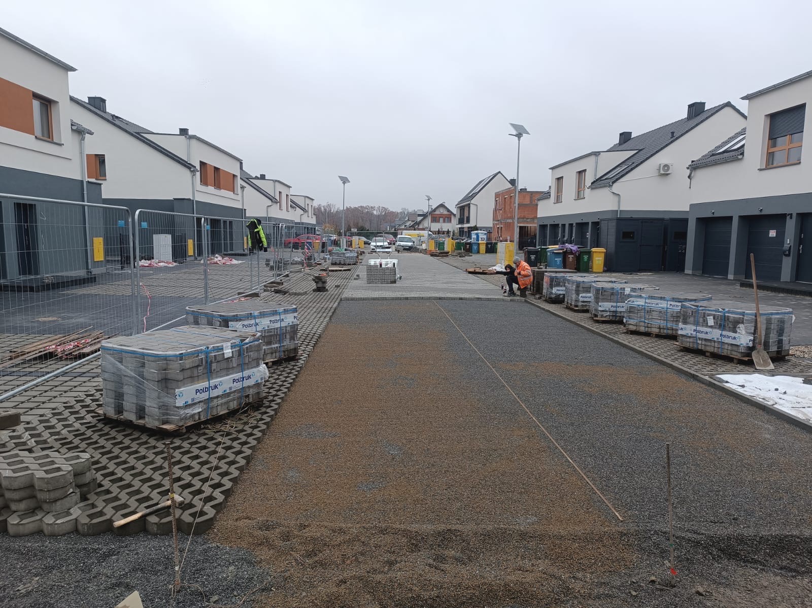 Plac budowy osiedla z widocznymi etapami przygotowania podłoża pod nawierzchnię, paletami kostki brukowej Polbruk i pracownikami w odblaskowych kamizelkach.
