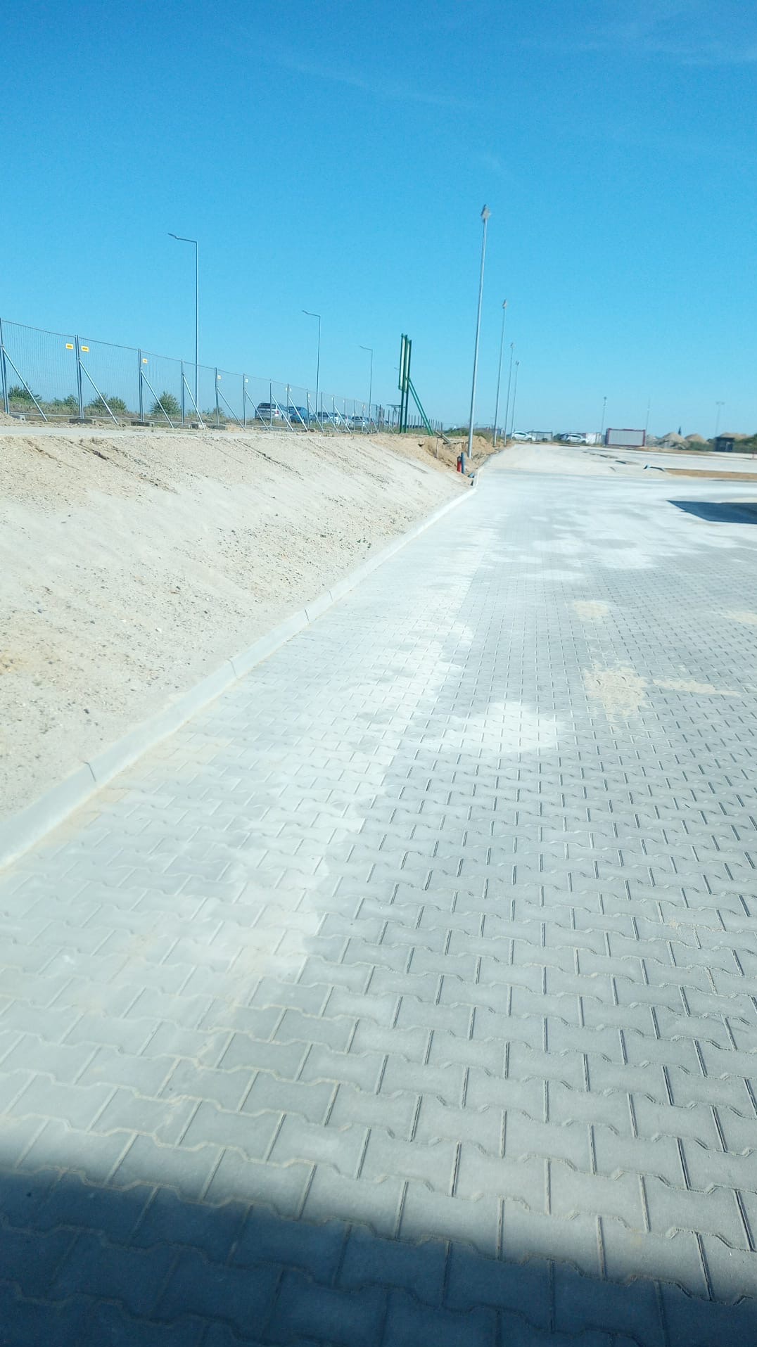 Nowo ułożona, szara kostka brukowa w przestrzeni przemysłowej, widoczna krawędź chodnika i ogrodzenie w tle.