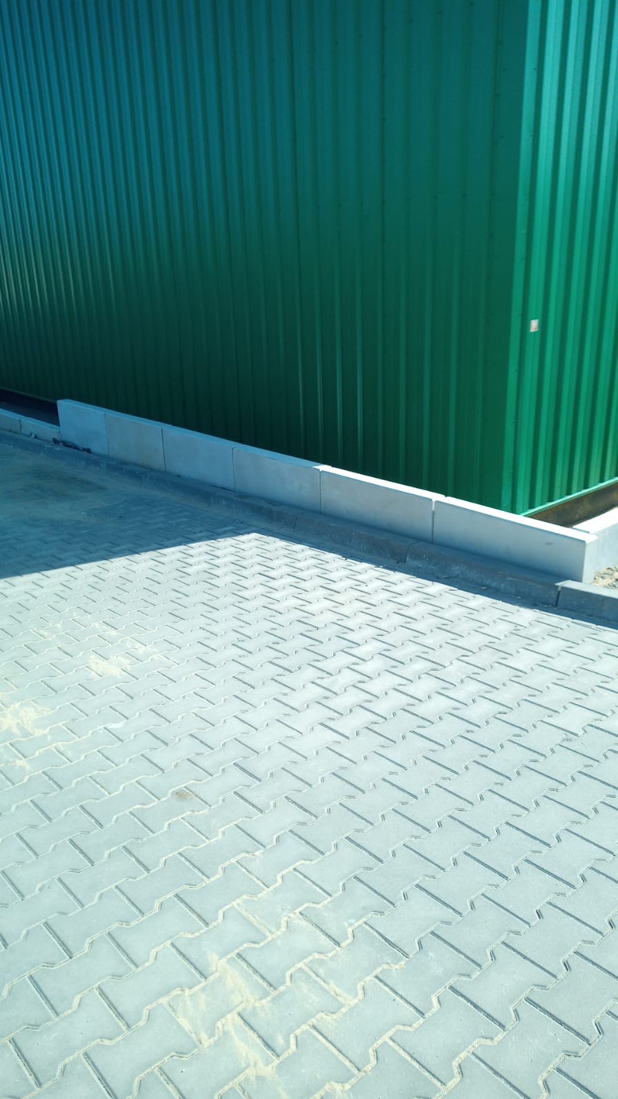 Ułożona szara kostka brukowa typu 'I' z betonowym obrzeżem i zieloną blachą trapezową w tle.