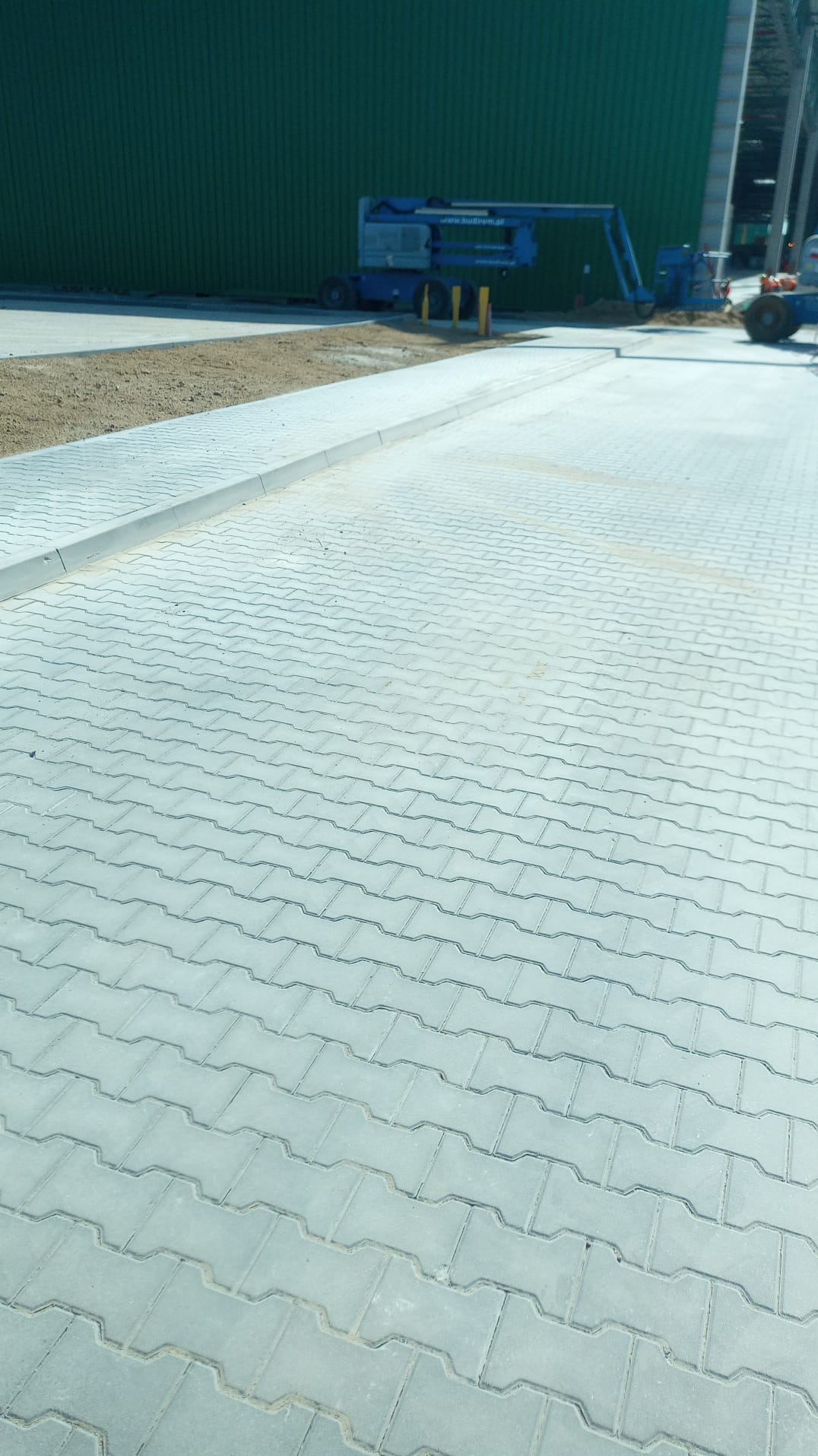 Ułożona szara kostka brukowa w jodełkę na placu budowy, w tle zielona hala i podnośnik koszowy.