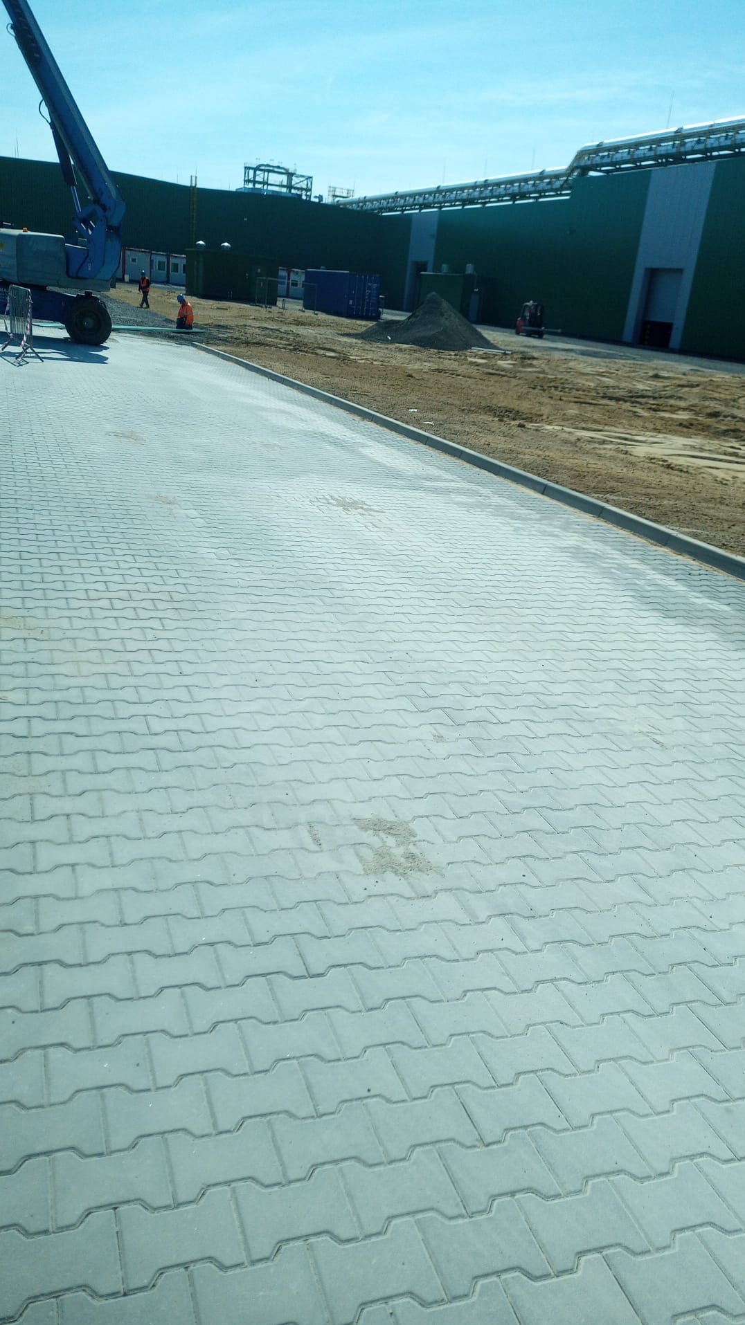Ułożona szara kostka brukowa w kształcie fali na terenie przemysłowym, w tle budynek magazynowy, maszyna budowlana i pracownicy w pomarańczowych kamizelkach.