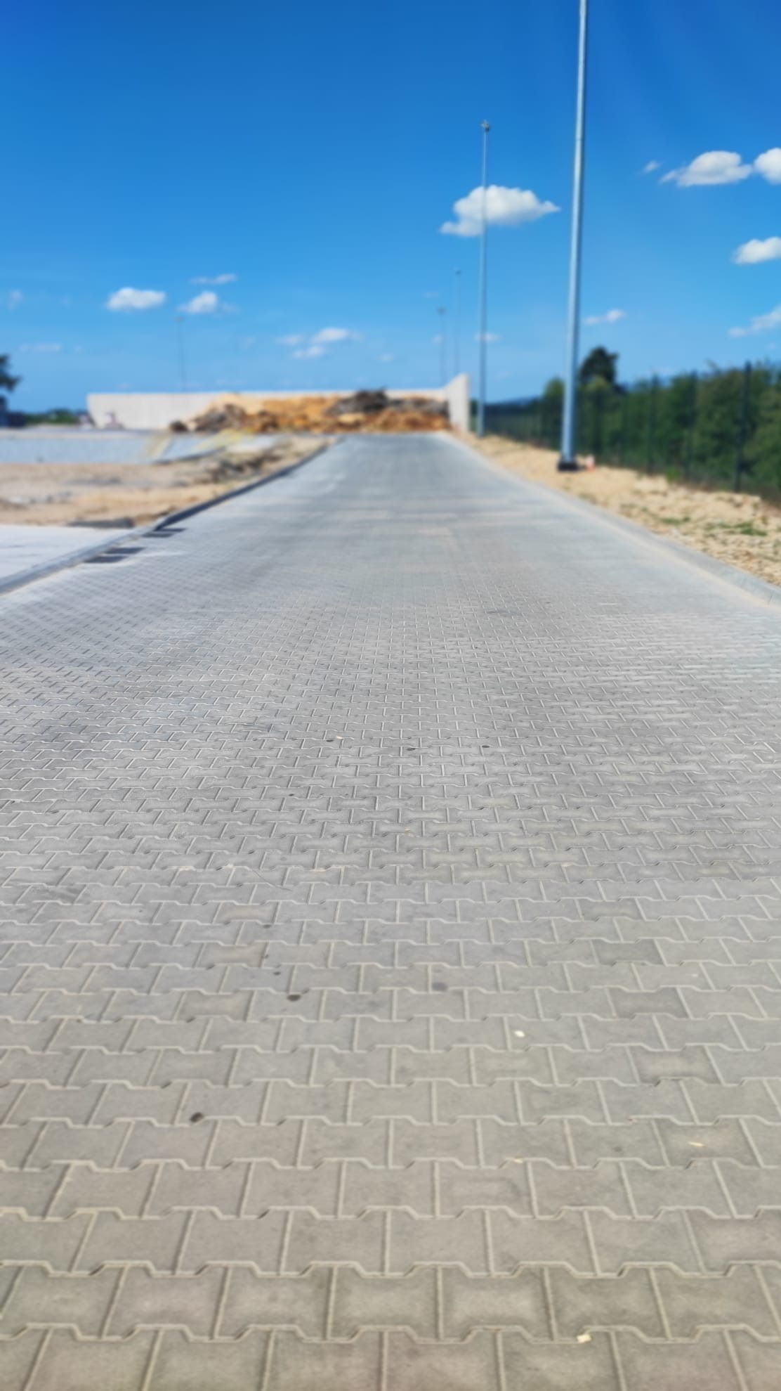Nowo ułożona, szara kostka brukowa w kształcie litery H tworzy długą ścieżkę w kierunku placu budowy pod błękitnym niebem.