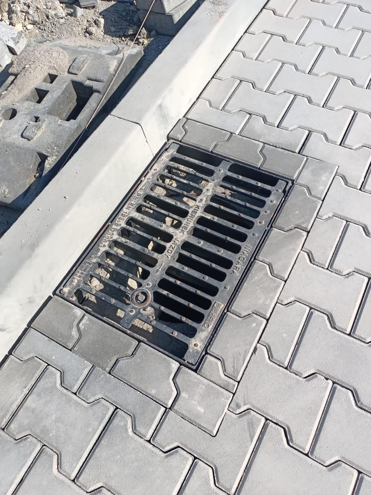 Szara kostka brukowa typu I ułożona wokół czarnej kratki ściekowej, widoczne krawężniki i przygotowane podłoże z piasku.