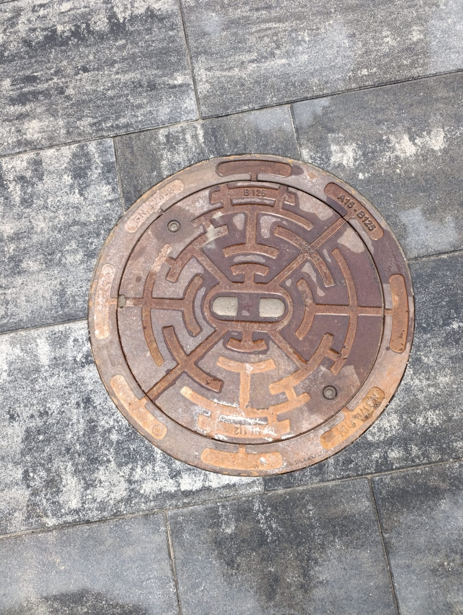 Szare kostki brukowe z okrągłą, rdzawą studzienką kanalizacyjną z labiryntowym wzorem pośrodku. Widok z góry, oświetlenie naturalne.