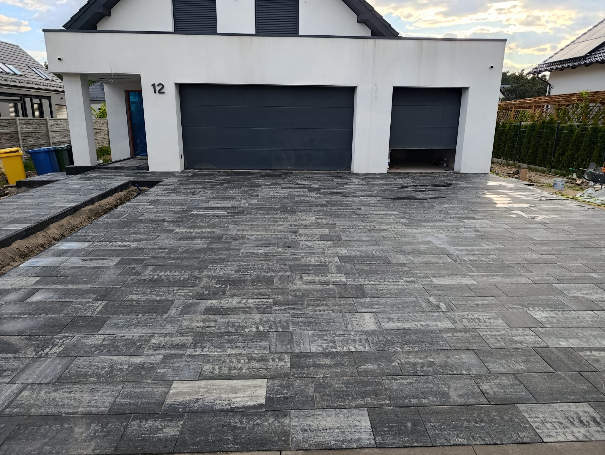 Ułożona kostka brukowa w odcieniach szarości przed nowoczesnym domem z numerem 12. Częściowo otwarty garaż. Widoczne przygotowane obrzeża.