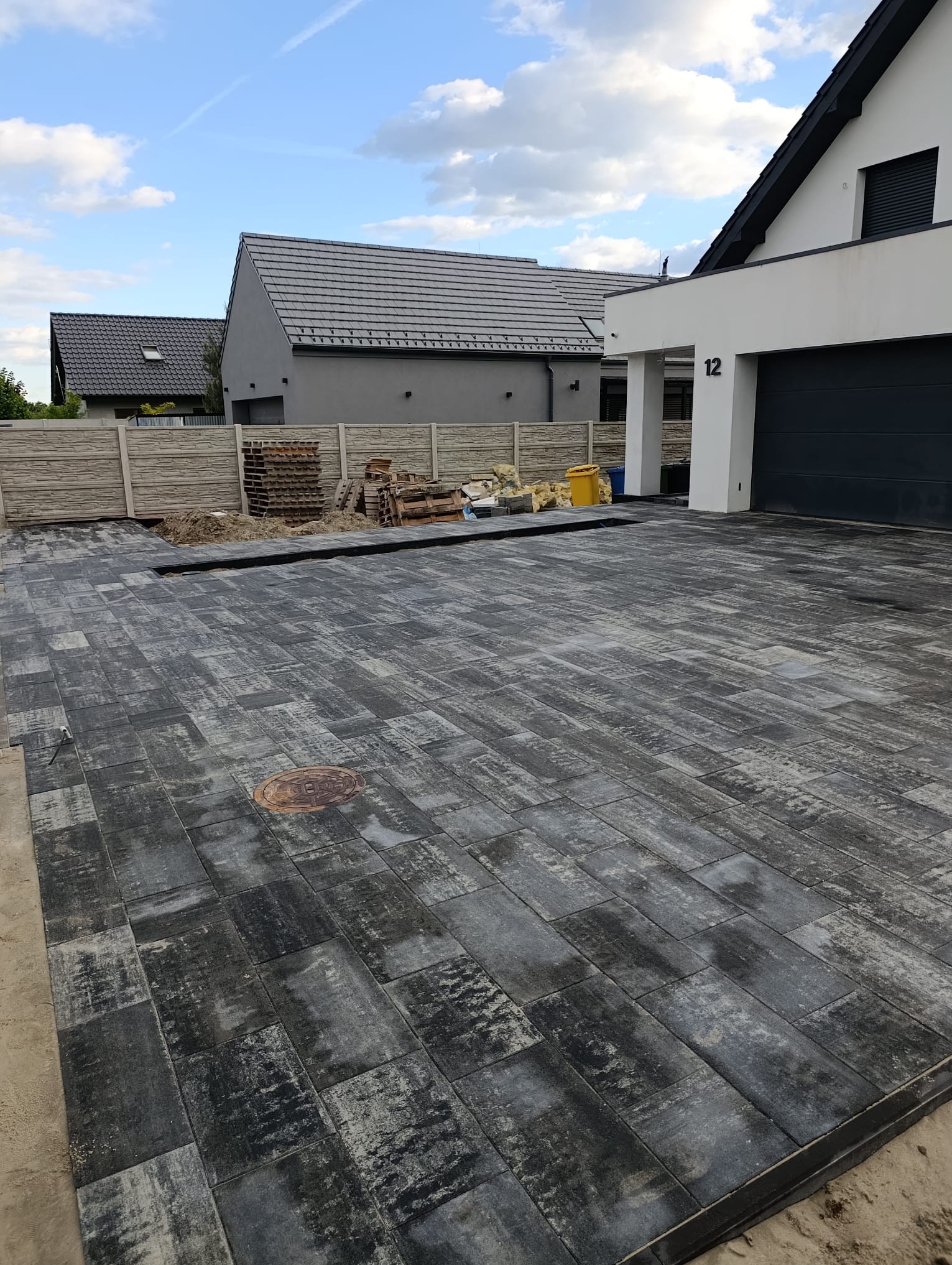 Nowoczesny podjazd z szarej, prostokątnej kostki brukowej, z widocznym włazem kanalizacyjnym i paletami kostki w tle, przy domu z numerem 12.