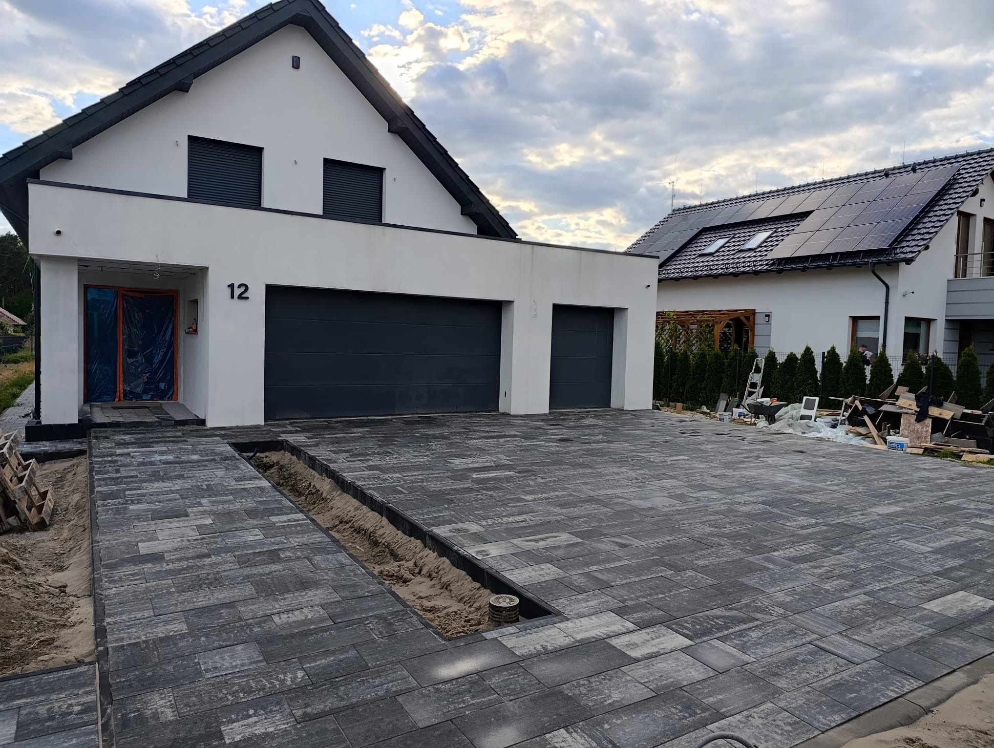 Częściowo ułożona, szara kostka brukowa na podjeździe przed nowoczesnym domem z dwoma garażami i numerem 12. Widoczny fragment odwodnienia liniowego.