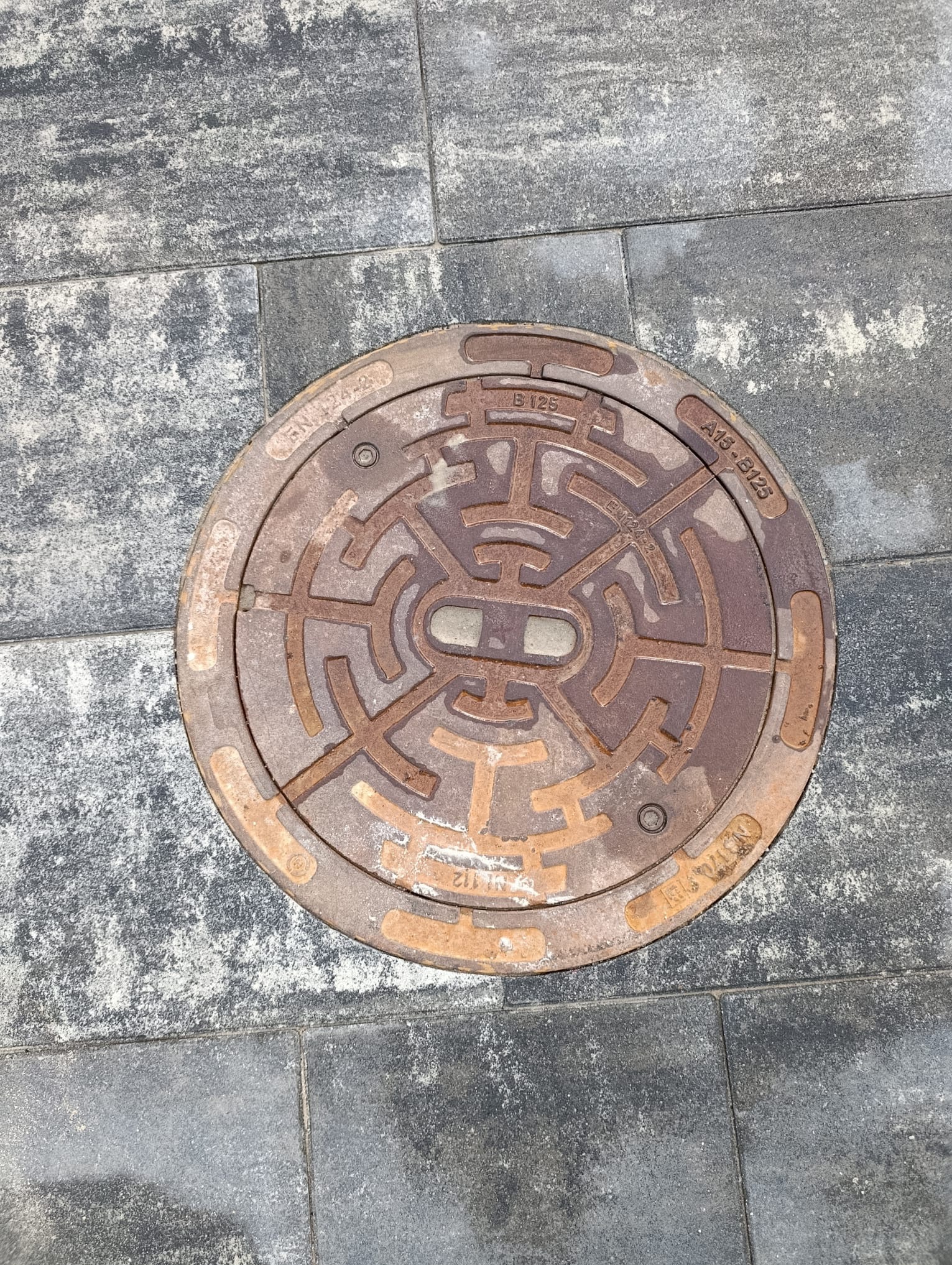 Szara kostka brukowa z okrągłą, żeliwną studzienką kanalizacyjną z labiryntowym wzorem, widoczne oznaczenia normy EN124-2. Detal nawierzchni.