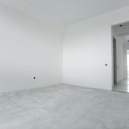 TOP NAT - Pusty, jasny pokój z surowymi ścianami i betonową podłogą, widoczne otwory na gniazdka elektryczne i przejście do kolejnych pomieszczeń w głębi.