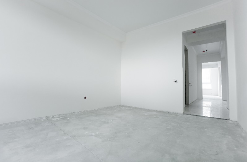 Pusty, jasny pokój z surowymi ścianami i betonową podłogą, widoczne otwory na gniazdka elektryczne i przejście do kolejnych pomieszczeń w głębi.