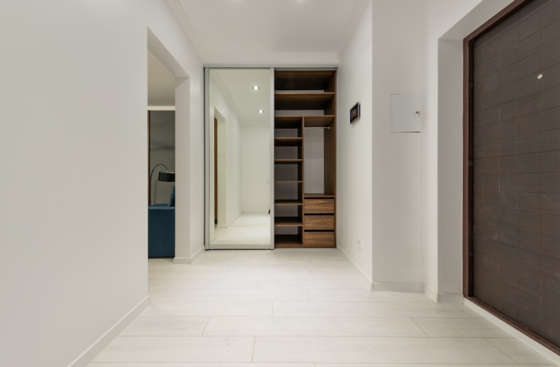 Nowoczesny przedpokój z białą podłogą, wbudowaną szafą z lustrzanymi drzwiami przesuwnymi i drewnianym regałem, oraz ciemnymi drzwiami wejściowymi po prawej stronie.