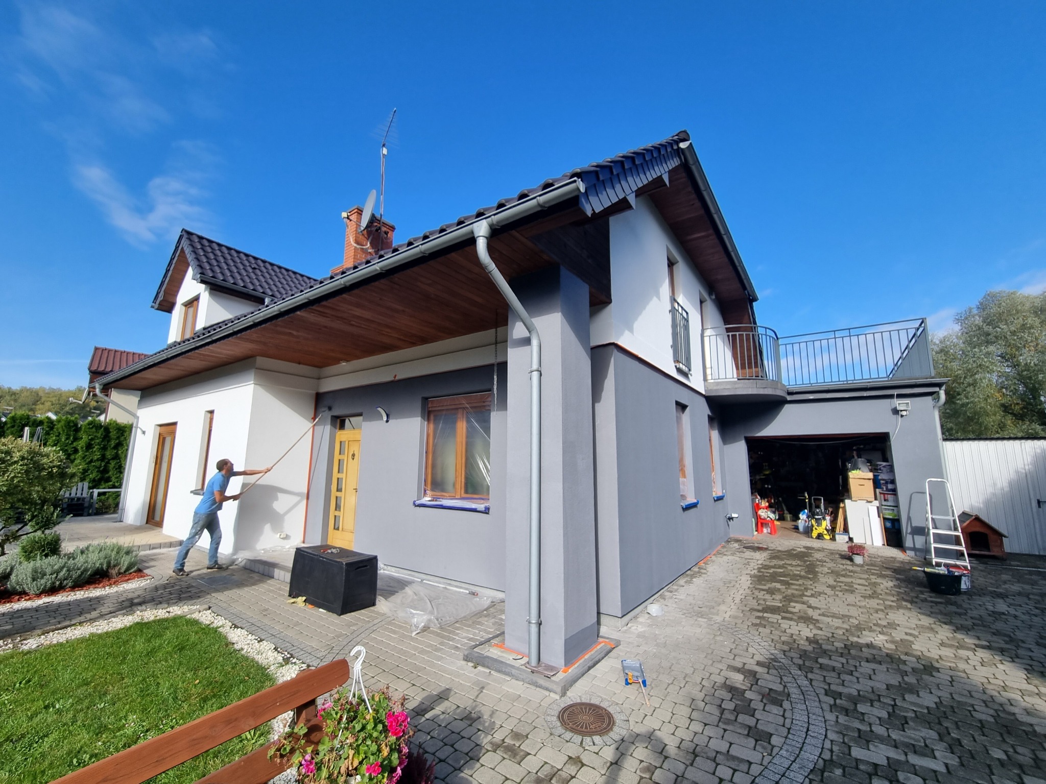 Mężczyzna maluje białą ścianę domu z szarą elewacją przy użyciu wałka na długim kiju, widoczna skrzynka narzędziowa i zabezpieczone okna folią malarską.