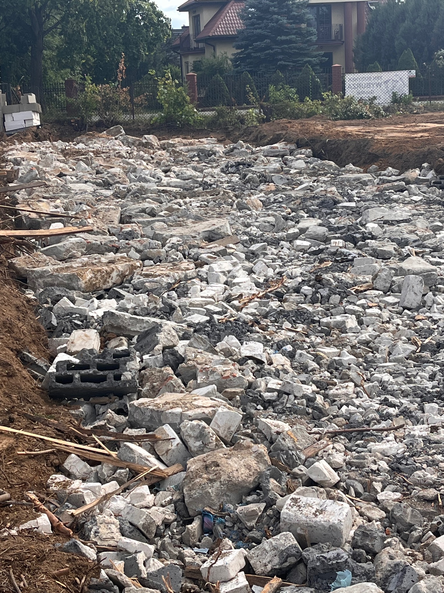 Gruzowisko po rozbiórce na działce budowlanej, z widocznym ziemnym wykopem i fragmentami domu w tle, w słoneczny dzień.