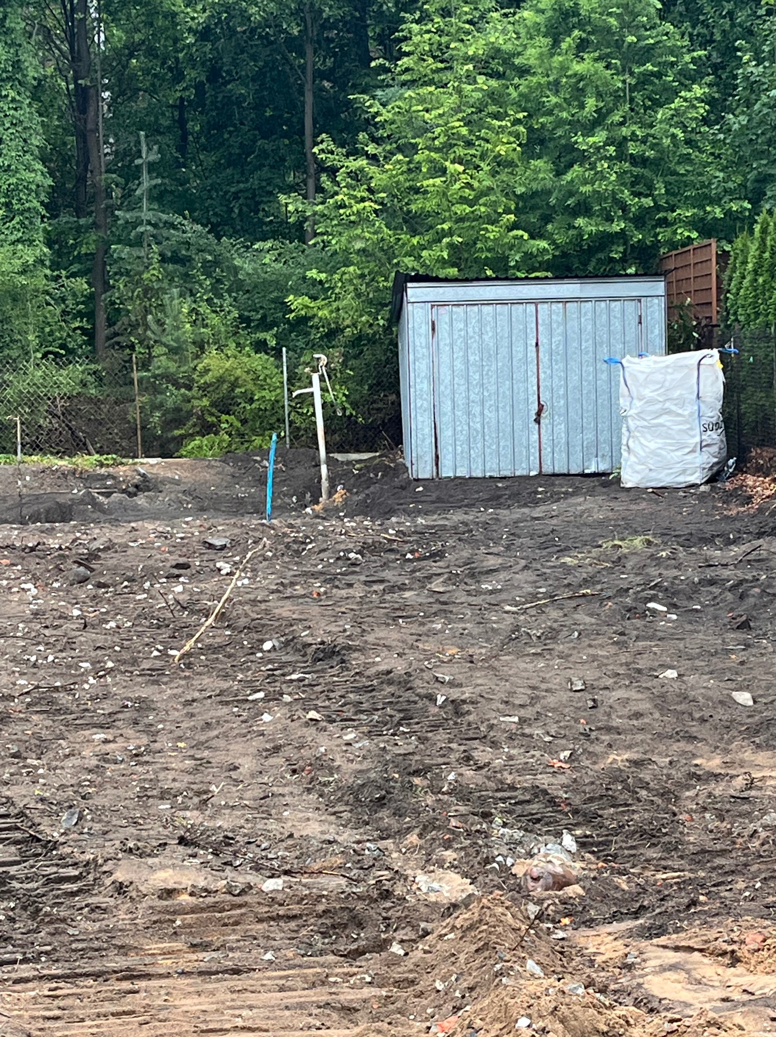 Teren po wyburzeniu z metalowym schowkiem i workiem. Widoczne ślady opon i gruz. W tle drzewa i ogrodzenie.