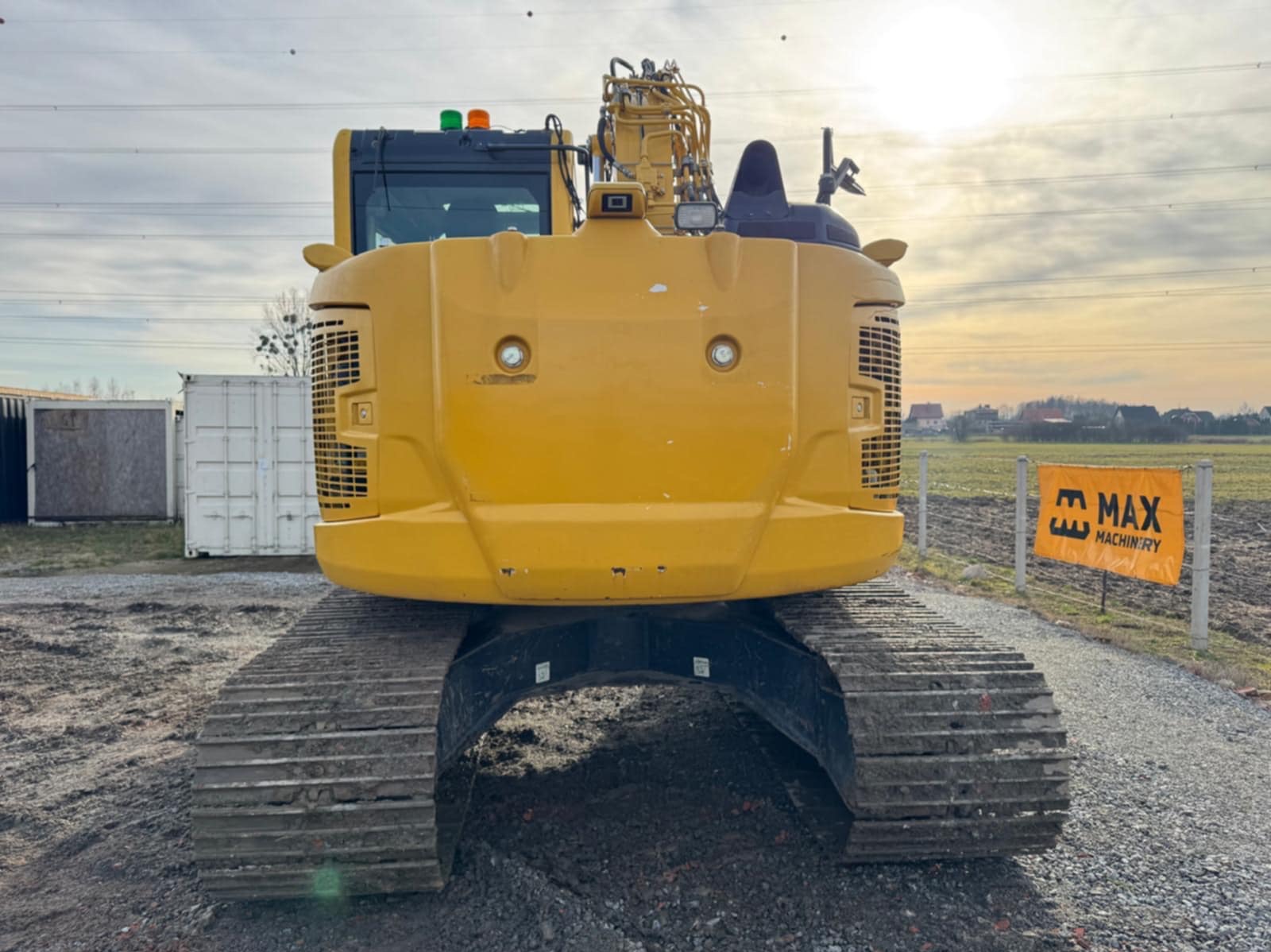 Żółta koparka gąsienicowa widziana z tyłu, na placu budowy, obok kontenerów i baneru reklamowego. Widoczne detale konstrukcyjne i oświetlenie maszyny.
