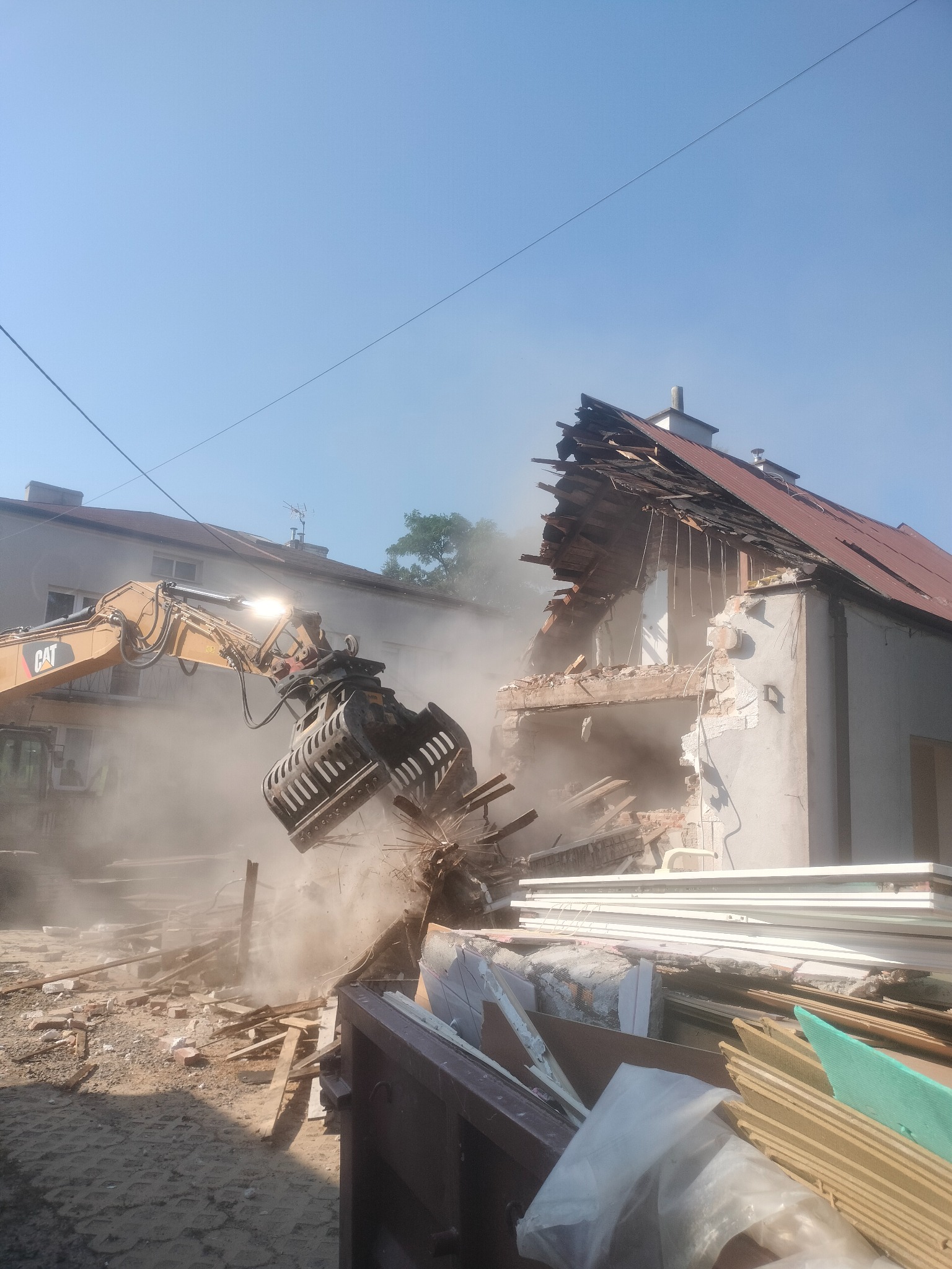 Żółty koparko-ładowarka CAT w trakcie rozbiórki budynku. Widoczne zniszczenia, gruz i pył. Na pierwszym planie składowane odpady. Jasne niebo.