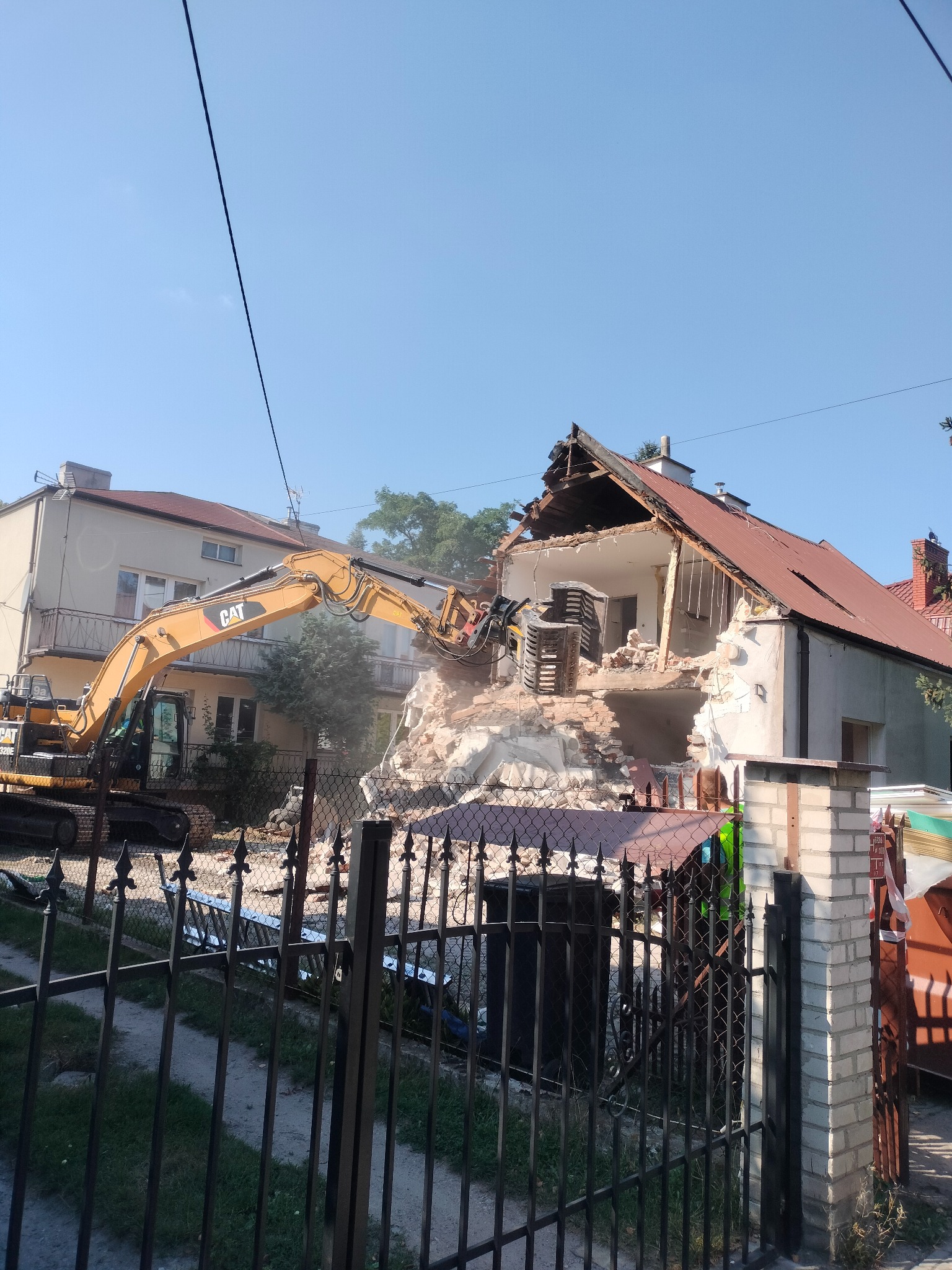 Żółta koparka CAT w trakcie rozbiórki domu z czerwonym dachem. Widok zza metalowego ogrodzenia, w tle budynki mieszkalne.