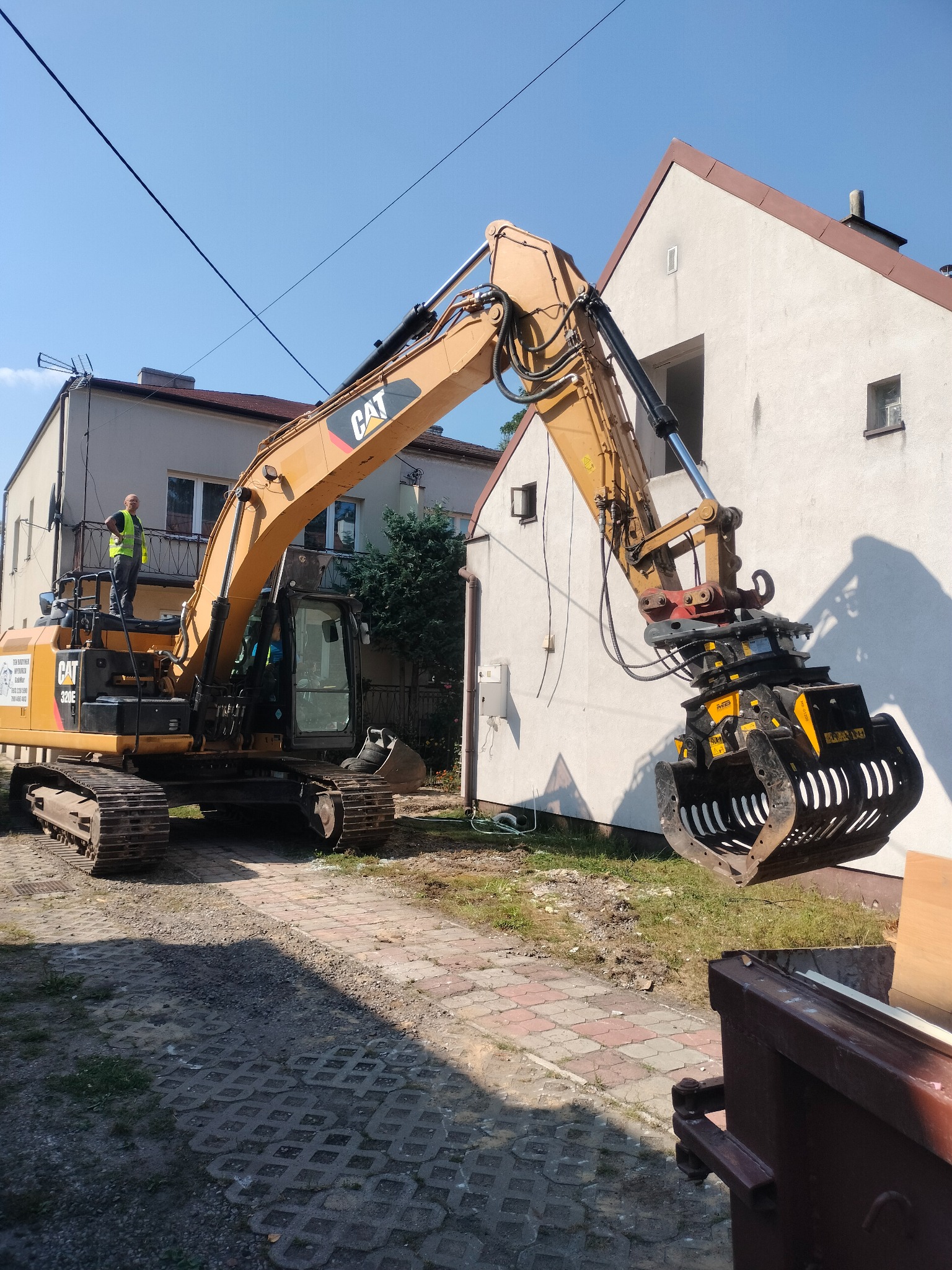Żółta koparka CAT 320E z chwytakiem MB w trakcie pracy przy budynku z białym tynkiem. Operator w kamizelce na maszynie. Słoneczny dzień, widoczne detale otoczenia.