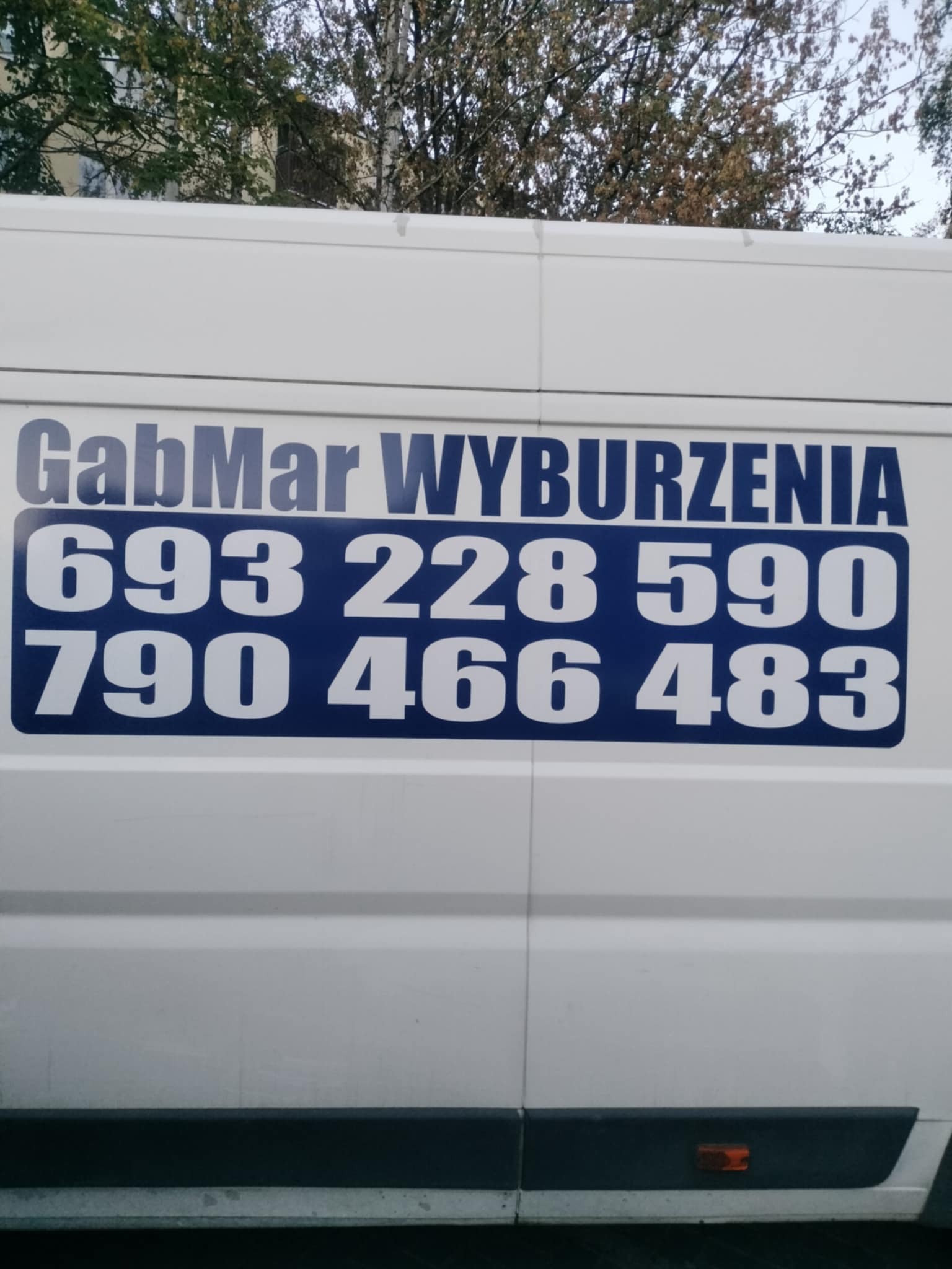 Biały samochód dostawczy z niebieskim napisem 'GabMar Wyburzenia' i numerami telefonów. Ujęcie z boku pojazdu.