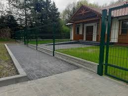 Ułożona kostka brukowa w odcieniach szarości prowadząca do domu z zielonym ogrodzeniem panelowym z podmurówką betonową. Widoczny fragment furtki.