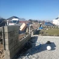 Wznoszenie muru z szarych bloczków betonowych na posesji, widoczne wystające zbrojenie, rozsypany żwir i zwinięte kable, biały samochód dostawczy w tle, błękitne niebo.