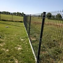 Długi, prosty odcinek nowoczesnego ogrodzenia z paneli zgrzewanych, widoczny z boku, z betonową podmurówką, na tle zielonej łąki i drzew w oddali, w słoneczny dzień.