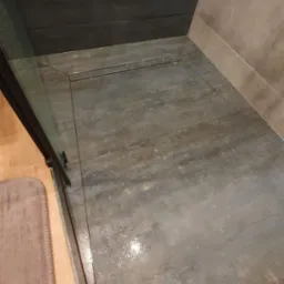 Nowoczesna kabina prysznicowa walk-in z szarymi płytkami imitującymi beton, odpływ liniowy i fragmentem szklanej ściany z czarnymi okuciami.