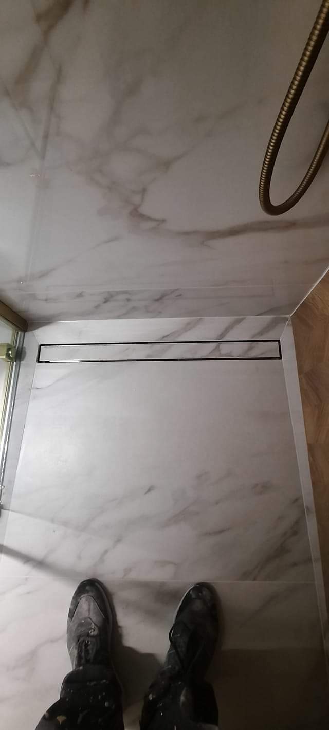 Wykończony brodzik prysznicowy z odpływem liniowym i marmurowymi płytkami, widok z góry na buty robocze.