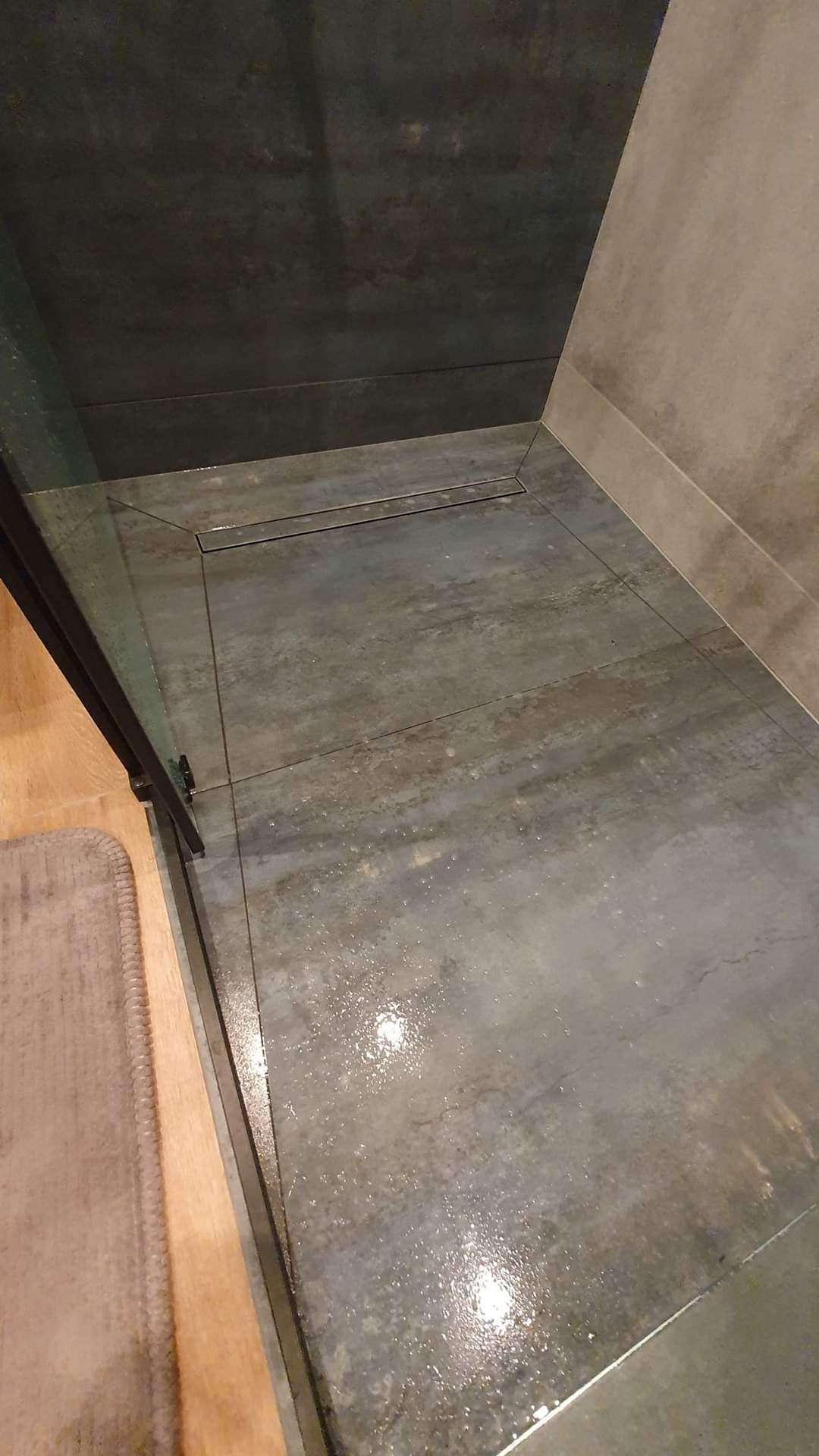 Nowoczesna kabina prysznicowa walk-in z szarymi płytkami imitującymi beton, odpływ liniowy i fragmentem szklanej ściany z czarnymi okuciami.