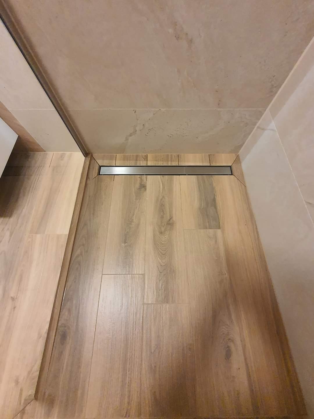 Wykończona łazienka z drewnopodobnymi panelami podłogowymi, beżowymi płytkami ściennymi i liniowym odpływem prysznicowym ze stali nierdzewnej.