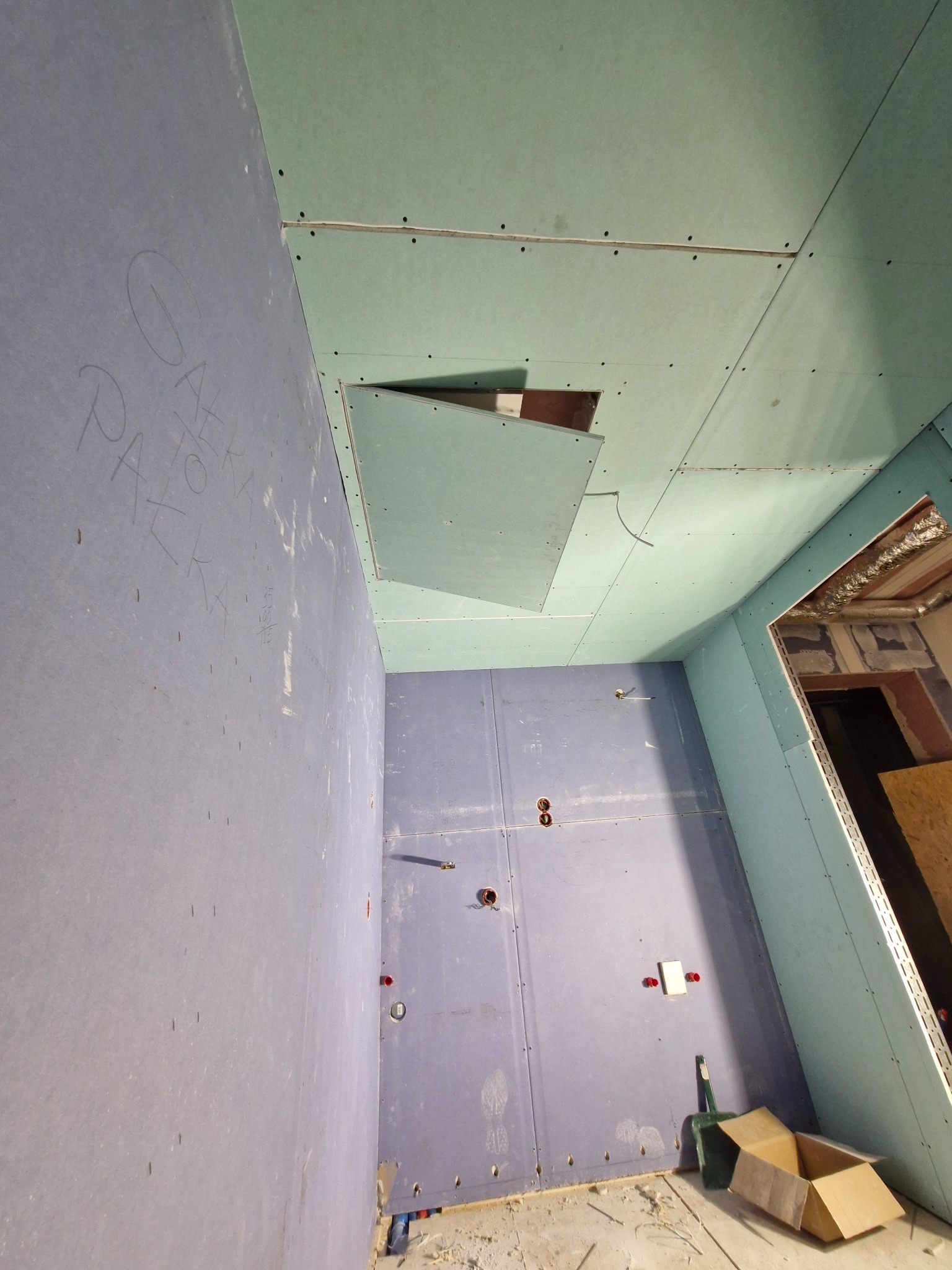 Wnętrze w trakcie prac wykończeniowych, ściany i sufit obłożone zielonymi i fioletowymi płytami, widoczne otwory instalacyjne i inspekcyjne, na ścianie napisy ołówkiem, na podłodze śmieci i karton.