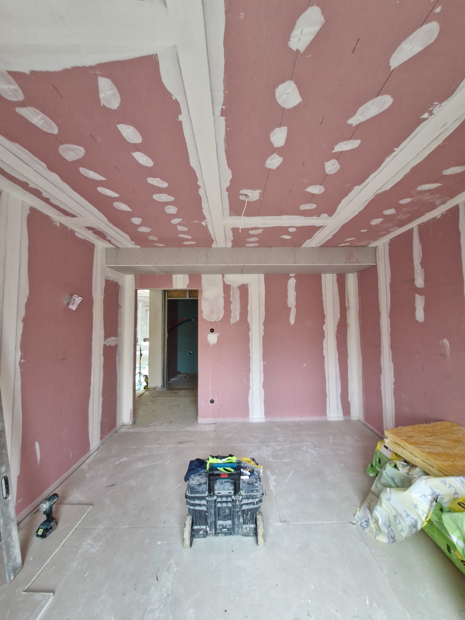 Wnętrze w trakcie montażu okładzin ściennych i sufitowych z płyt gipsowo-kartonowych, widoczna skrzynka z narzędziami, szpachlowane łączenia płyt i otwór drzwiowy w tle.