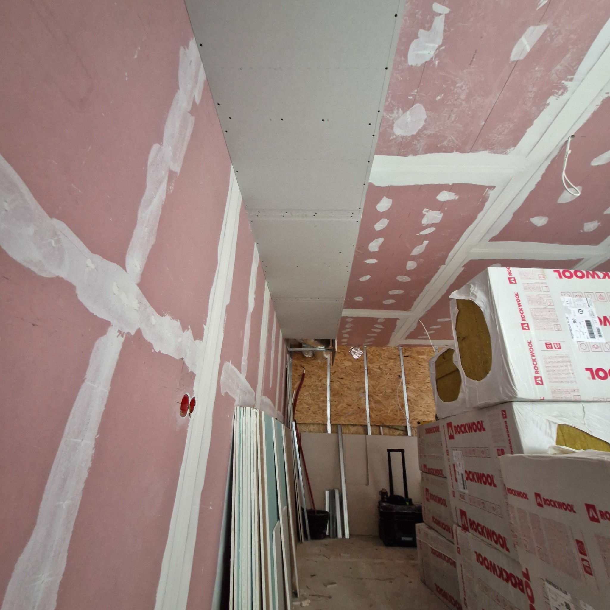 Wnętrze w trakcie prac wykończeniowych: ściany pokryte różową farbą z widocznymi łączeniami, częściowo zamontowane płyty na suficie, paczki wełny mineralnej Rockwool oraz stos płyt w rogu...