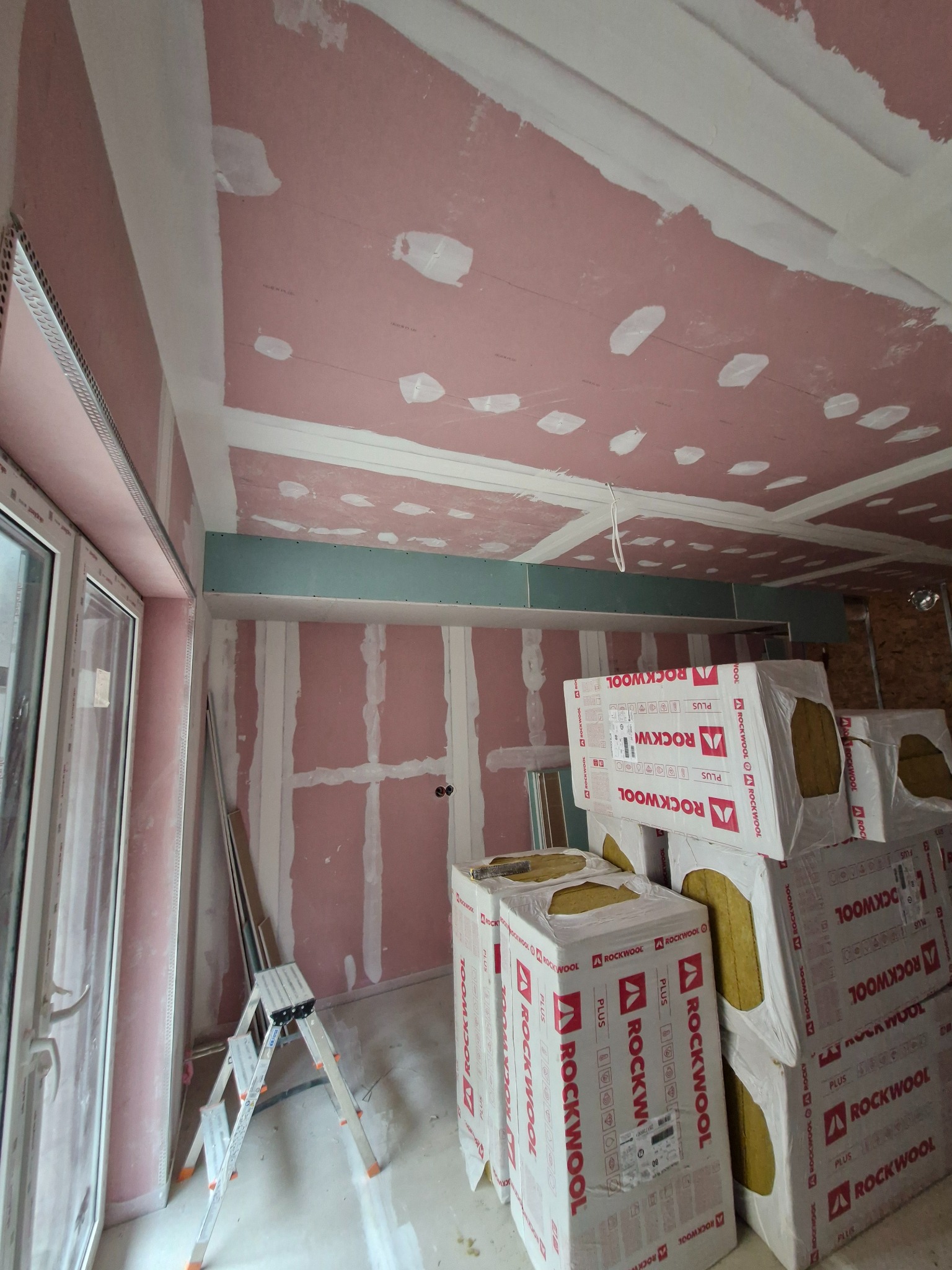Wnętrze w trakcie remontu, widoczne ściany i sufit pokryte różową warstwą, częściowo zaszpachlowane, obok okna stoi drabina, a w głębi pomieszczenia składowane paczki wełny mineralnej Rockwool.